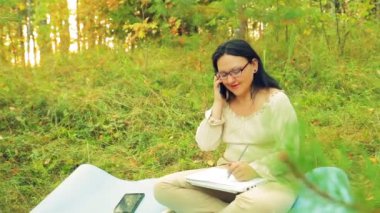 Bir orman içinde genç bir kadın bir defterde yazılı ve cep telefonu üzerinde konuşmaya çalışır