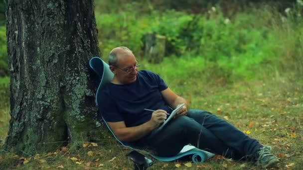 Человек в очках сидит под деревом в парке и пишет в блокноте карандашом — стоковое видео