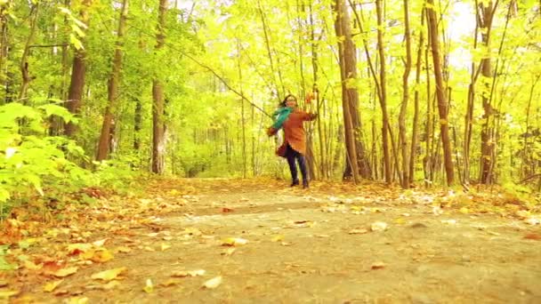 Een jonge brunette vrouw in een jas en sjaal op een zonnige dag in een herfst park met esdoorn bladeren in haar handen loopt langs een pad en gooit omhoog bladeren — Stockvideo