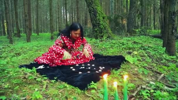 Una joven gitana en el bosque realiza movimientos mágicos con las manos sobre velas encendidas y guijarros blancos . — Vídeos de Stock