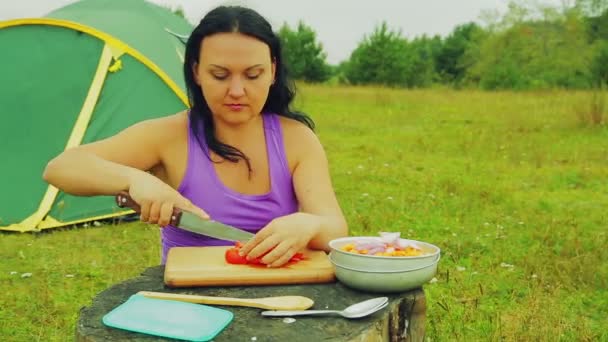 Eine junge Frau in der Nähe eines Zeltes schneidet rote Tomatenscheiben auf einem Holzbrett. — Stockvideo