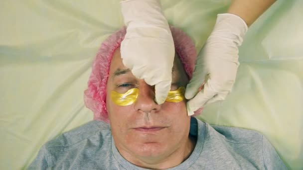Las manos de una mujer esteticista en guantes se retiran de la cara de las almohadillas de colágeno hombre debajo de los ojos — Vídeos de Stock