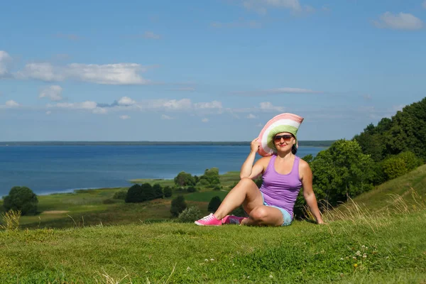 Mujer joven viajera en sombrero y gafas de sol descansando en el borde de una colina frente a un lago . —  Fotos de Stock
