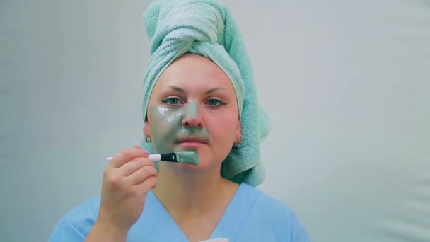 A young woman after a shower applies a mud mask on her face with a brush. Looking at the camera. — Stock Video