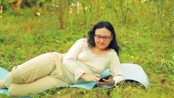 A young cheerful woman in glasses in the forest lies on a rug for yoga and communicates in a messenger — Stock Video