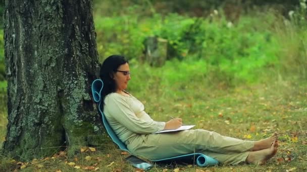 Een vrouw in glazen barefoot zit onder een boom in het park en een potlood tekent in een notitieblok — Stockvideo