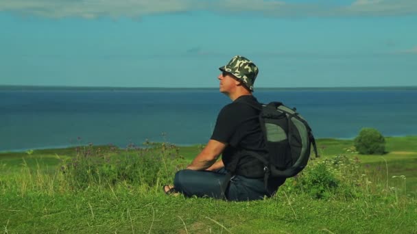 Un hombre con una mochila detrás de su espalda se sienta en el borde de la montaña y admira la vista del lago. Disparo desde el costado . — Vídeos de Stock