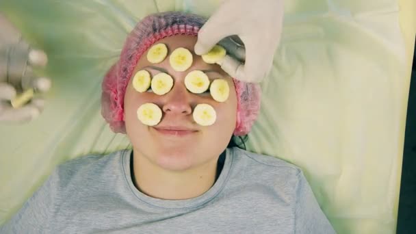 As mãos do homem do cosmetician retiram da cara da mulher uma máscara de uma banana. Vista de cima . — Vídeo de Stock