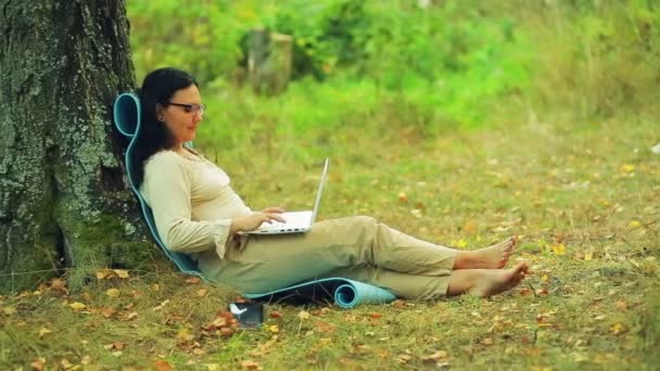 Una mujer en gafas descalza se sienta debajo de un árbol en el parque y trabaja con una computadora portátil . — Vídeos de Stock