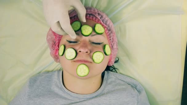 Handen van een mannelijke schoonheidsspecialist in handschoenen verwijderen uit de stukken van het vrouwelijke gezicht van komkommer — Stockvideo