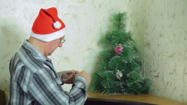 Un uomo in un cappello di Babbo Natale sta selezionando le palle di festa per decorare un albero di Natale — Video Stock