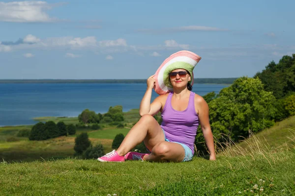 Una joven con sombrero y gafas de sol descansa en el borde de una colina frente a un lago . —  Fotos de Stock
