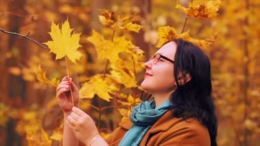 Sonbahar parkta gözlüklü genç gülümseyen kadın desenli akçaağaç yaprağı elinde tutar ve bunu inceliyor.