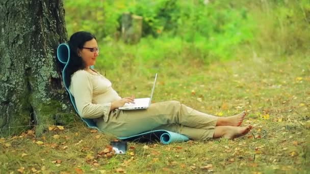 Женщина в очках сидит спиной к дереву в парке и работает с ноутбуком — стоковое видео