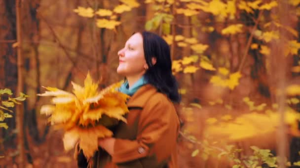 Giovane donna bruna sorridente nel parco autunnale sta girando con foglie d'acero nelle sue mani — Video Stock