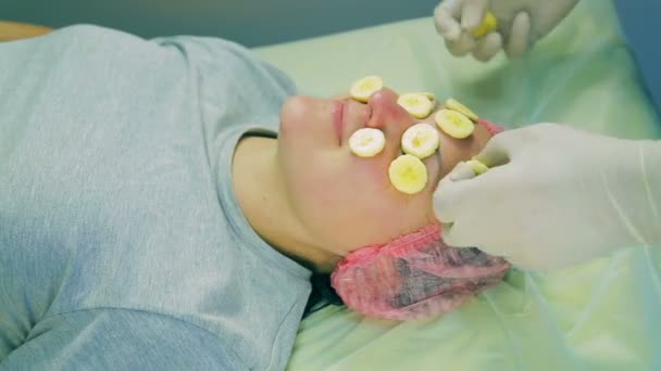 As mãos de um cosmetologist masculino em luvas retiram partes de uma banana da cara de mulheres — Vídeo de Stock
