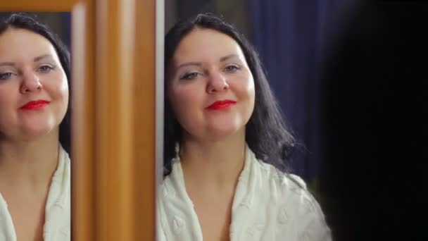 A young woman in a white robe in front of a mirror with bright red lipstick is smiling and flirting with her lips. — Stock Video