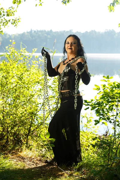 A young witch in black clothes with chains in his hands against the background of the lake. Vertical photo. — Stock Photo, Image