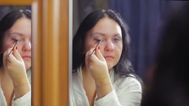 Een jonge vrouw in een witte jas voor een spiegel schildert haar ogen met schaduwen met een borstel — Stockvideo