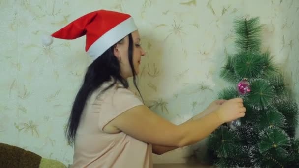 En kvinna som ler i Santas hatt sätter en holiday boll på julgranen — Stockvideo