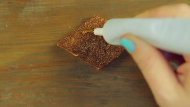 Una mano sostiene un tubo de pintura para cocinar y dibuja caras divertidas en las galletas de chocolate con pintura amarilla — Vídeo de stock