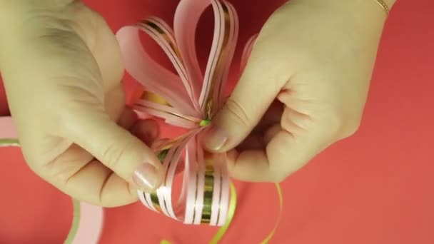 As mãos das mulheres endireitam as pétalas de um arco de presente de fita rosa e dão-lhe a forma de um arco. Fundo vermelho — Vídeo de Stock