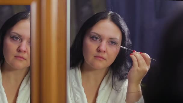 Een jonge vrouw in een witte jas voor een spiegel schildert haar ogen met schaduwen met een borstel — Stockvideo