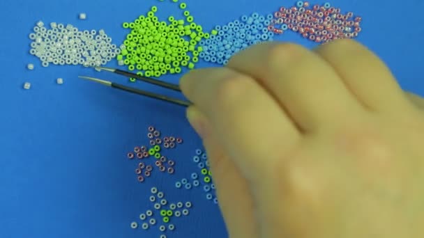 Női kéz a kék háttér bökések ki több színű gyöngyöket virágai, csipesszel. Közeli kép: — Stock videók