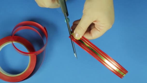 Le mani femminili tagliano gli angoli con un paio di forbici per un fiocco regalo nastro rosso. Fondo blu — Video Stock
