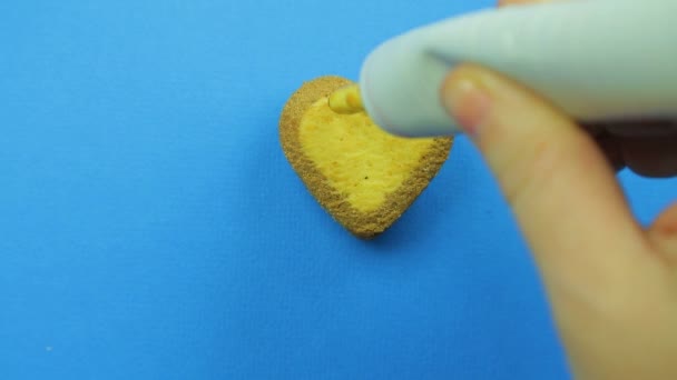 A mão feminina desenha emoticons engraçados em biscoitos em forma de coração em um fundo azul com cobertura marrom de um tubo — Vídeo de Stock