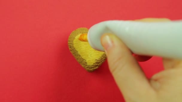 A mão feminina pinta biscoitos em forma de coração em um fundo vermelho com um esmalte de caramelo de um tubo caras engraçadas — Vídeo de Stock