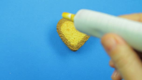 Una mano femenina dibuja caras graciosas sobre un fondo azul con un glaseado marrón de un tubo sobre una galleta en forma de corazón sobre un fondo azul — Vídeo de stock