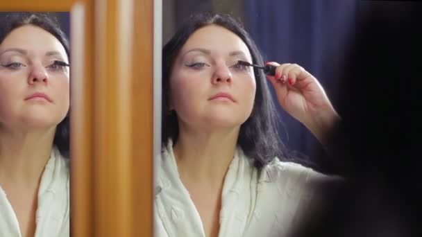 Una joven con un abrigo blanco frente a un espejo se pinta las pestañas con rímel — Vídeos de Stock