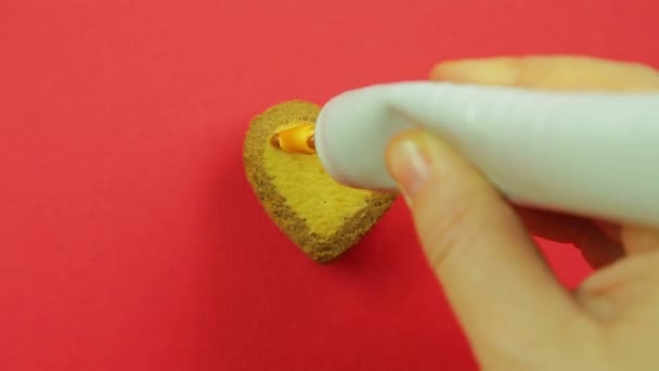 Ženská ruka maluje sušenky ve tvaru srdce na červeném pozadí s karamelovou polevou z trubice legrační obličeje — Stock video