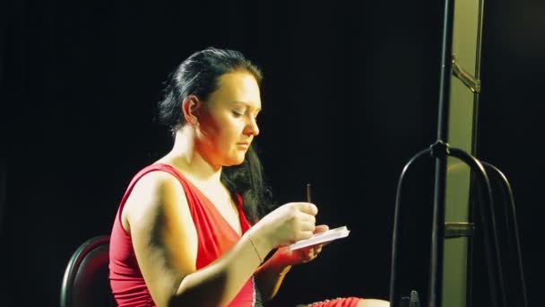 Jonge vrouw in een rode jurk voor een spiegel met een borstel zet schaduw op de oogleden — Stockvideo