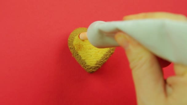 Γυναικείο χέρι αντλεί ένα μπισκότο σε σχήμα καρδιάς σε κόκκινο φόντο με κόκκινο σωλήνα ζαχαροαλοιφή αστείες φατσούλες. Χειροποίητο — Αρχείο Βίντεο
