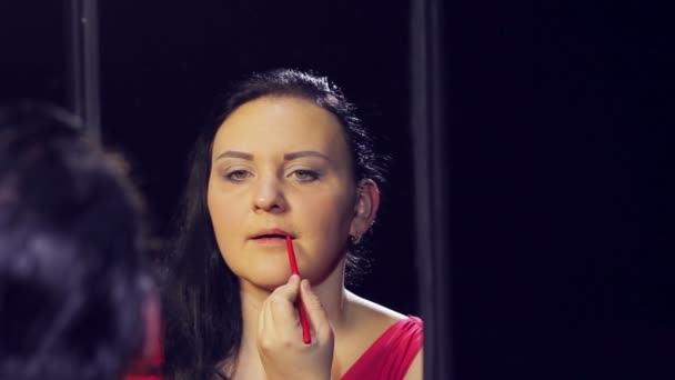 A young brunette woman in a red dress in front of a mirror applies a red contour to her lips. — Stock Video