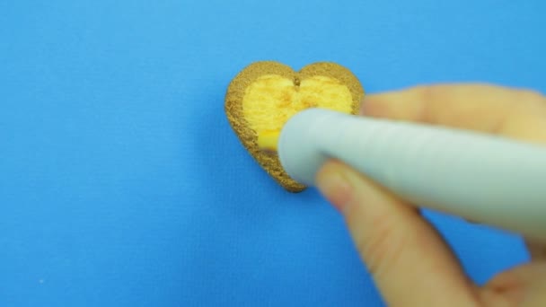 Weibliche Hand zeichnet lustige Emoticons auf herzförmige Kekse auf blauem Hintergrund mit braunem Zuckerguss aus einer Röhre — Stockvideo