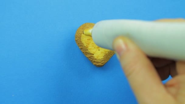 Een vrouwelijke hand trekt grappige gezichten op een blauwe achtergrond met een bruin suikerglazuur uit een buis op een hart-vormige cookie op een blauwe achtergrond — Stockvideo