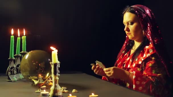 Μια νεαρή τσιγγάνα σε ένα κόκκινο φόρεμα σε ένα τραπέζι των κεριών διαβάζει το μέλλον σχετικά με τις κάρτες. Το σχέδιο των μέσων — Αρχείο Βίντεο
