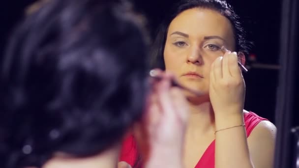 Una giovane donna bruna in un vestito rosso davanti a uno specchio mette ombre luminose sulle sue palpebre — Video Stock