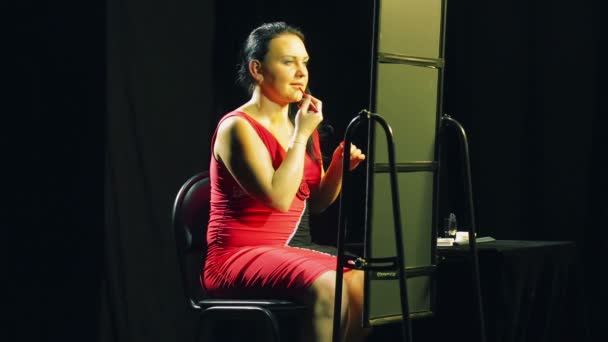 Een jonge vrouw in een rode jurk voor een spiegel zet een rode omtrek op haar lippen — Stockvideo
