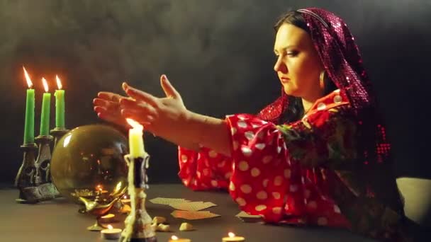 Молодой цыган в красном платье за столом при свечах читает будущее на волшебном шаре. Средний план — стоковое видео