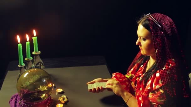 Молодой цыган в красном платье за столом при свечах читает будущее с карточками судьбы. Средний план . — стоковое видео