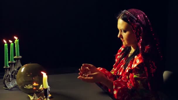 Молодой цыган в красном платье за столом при свечах бросает гальку в стол для гадания. Средний план — стоковое видео