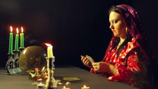 Um jovem cigano em um vestido vermelho em uma mesa à luz de velas lê o futuro nos cartões. O plano médio . — Vídeo de Stock