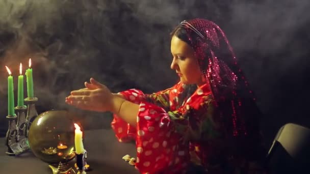 Um jovem cigano em um vestido vermelho em uma mesa à luz de velas lê o futuro em uma bola de vidro mágica em fumaça. O plano médio — Vídeo de Stock