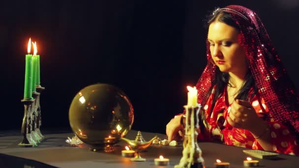 Μια νεαρή τσιγγάνα σε ένα κόκκινο φόρεμα σε ένα τραπέζι των κεριών διαβάζει το μέλλον πάνω από τις πέτρες. Το σχέδιο των μέσων — Αρχείο Βίντεο