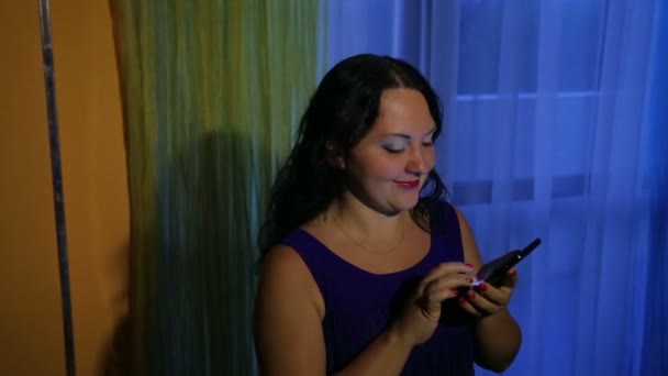 一位身穿蓝色衣服的年轻女子在窗户旁边在智能手机上留言 — 图库视频影像