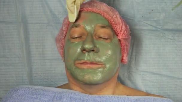 Eine Kosmetikerin in Handschuhen trägt mit einem Pinsel eine therapeutische Lehmmaske auf das Gesicht eines Mannes auf. Zeitrunden — Stockvideo